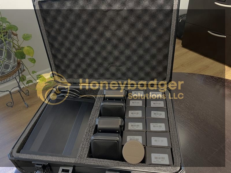 An open black case with foam holds labeled electronic devices, cables, and a round object on a dark wooden surface.