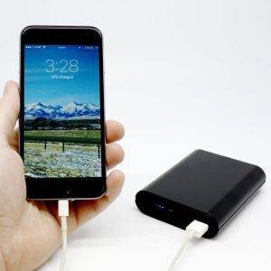 A hand holds a smartphone displaying a mountain wallpaper, connected to a black Power Bank via a white charging cable.