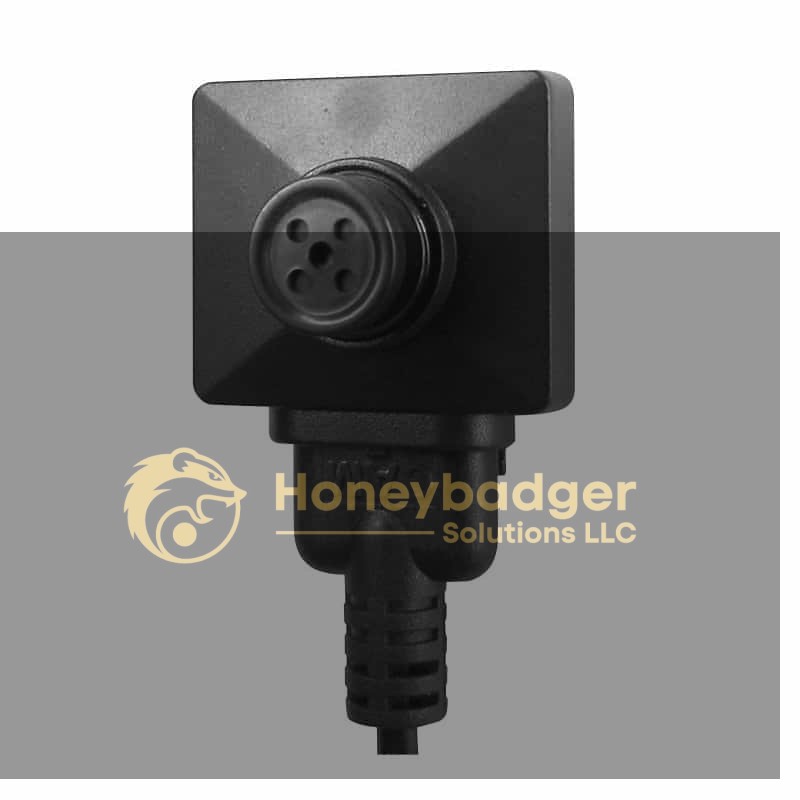 A compact black button camera with a square design and a circular lens, attached to a cable.