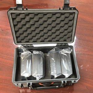 An open black hard-shell case with foam lining, containing two gray rectangular components wrapped in clear plastic.