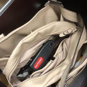 A beige handbag unzipped, showing a black device labeled 'Spy Finder Pro' among other items inside.