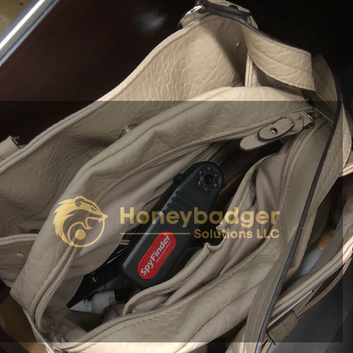 A beige handbag unzipped, showing a black device labeled 'Spy Finder Pro' among other items inside.