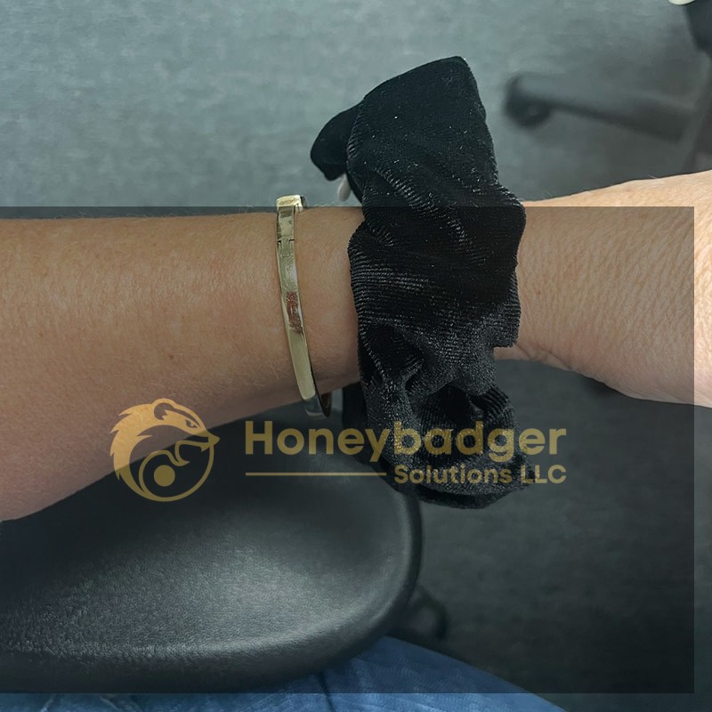 A wrist adorned with a shiny gold bracelet and a black velvet scrunchie, resting on a desk.