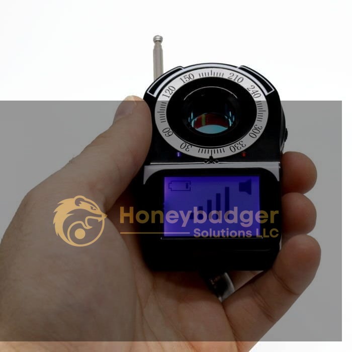 A hand holds a Camera Finder with RF Detector, a small black device with a circular dial and blue digital screen displaying battery and signal strength.