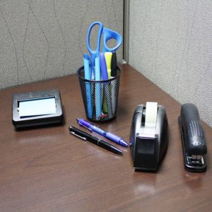 A black mesh pen holder containing scissors, pens, and highlighters on a brown desk with a tape dispenser and stapler.