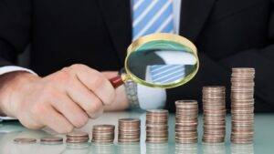 A person in a suit using a magnifying glass to examine stacks of coins arranged in increasing height.