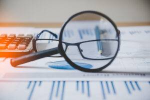 A magnifying glass and eyeglasses placed on financial documents with charts and graphs, alongside a calculator.