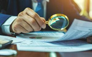 A person in a suit uses a magnifying glass to inspect financial documents and charts on a desk in an office setting.