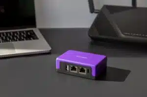 Firewalla Purple, a compact purple Gigabit Cyber Security Firewall and Router with multiple ports, placed on a dark table.