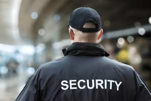 A security guard wearing a black jacket with 'SECURITY' in bold white letters stands in a public space.