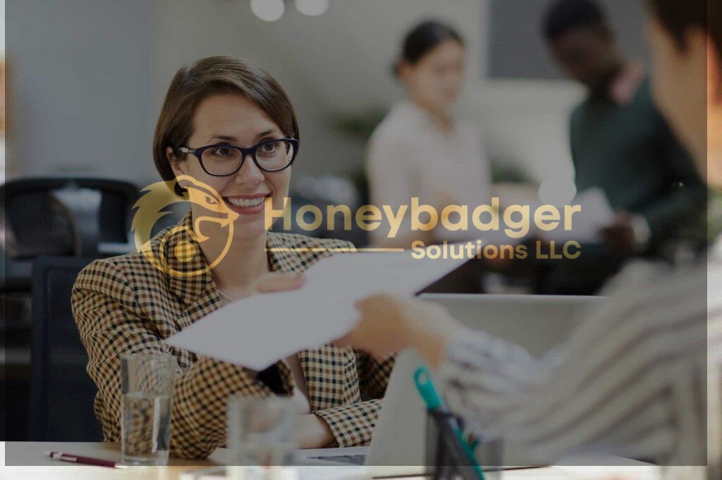 A smiling woman wearing glasses and a plaid blazer sits at a desk, receiving papers from another person in an office setting.
