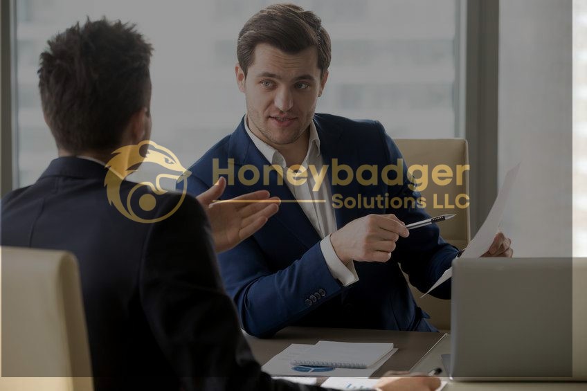 Two businessmen in suits seated at a table, one holding a document and gesturing while speaking to the other.