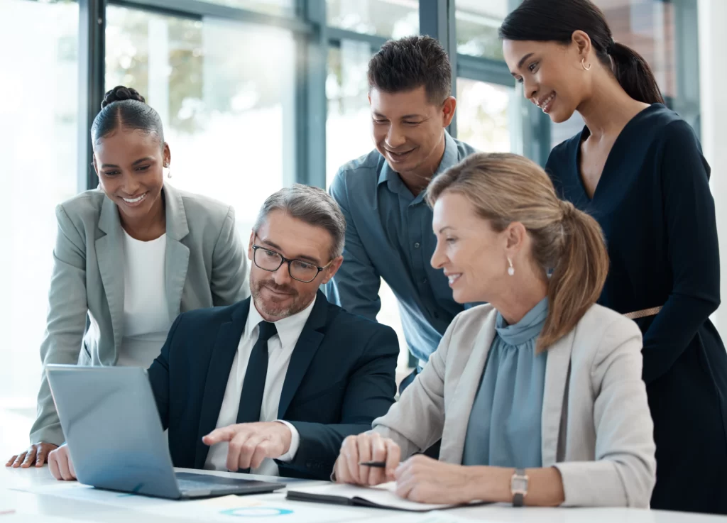 A group of five professionals engaged in a discussion while looking at a laptop screen in a modern office setting.