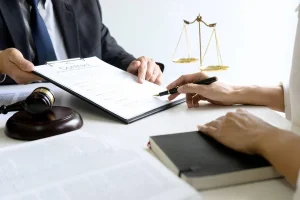 Two individuals at a desk, one presenting a clipboard while the other points with a pen. A gavel and legal scales are visible.