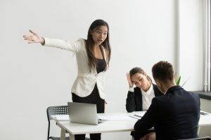 A woman in a white blazer stands with one arm extended, while two colleagues appear stressed at a table in an office setting.