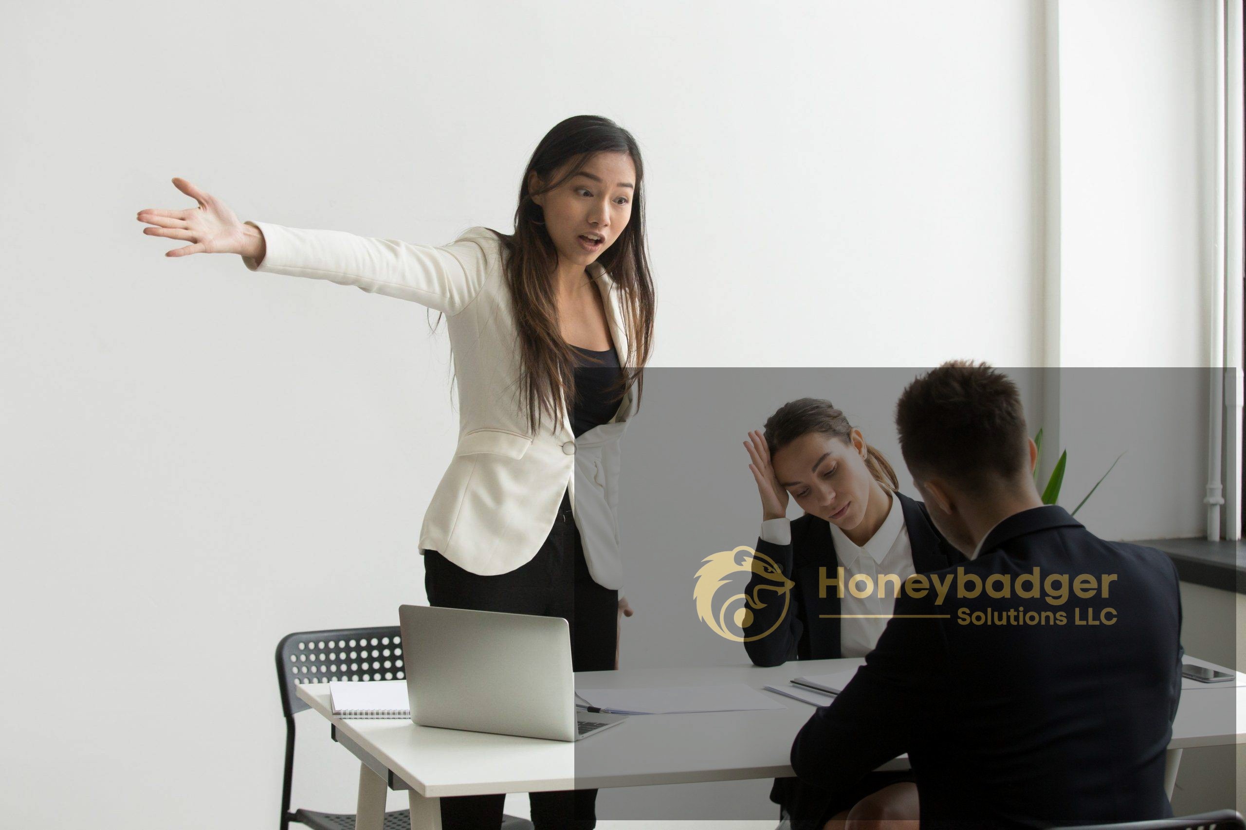 A woman in a white blazer stands with one arm extended, while two colleagues appear stressed at a table in an office setting.