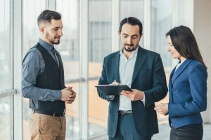 Three business professionals in formal attire discussing strategies while one man writes on a clipboard.