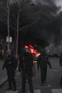 Riot police in protective gear standing in front of a burning vehicle with thick black smoke rising.
