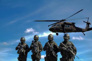 Four armed soldiers in black tactical gear stand in formation with a military helicopter flying overhead against a blue sky.