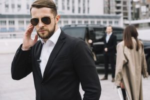 A man in a black suit and sunglasses stands in the foreground, touching his earpiece, with a black SUV in the background.