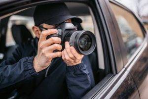 A person in a dark jacket and cap holds a camera, taking photos through the open window of a car.
