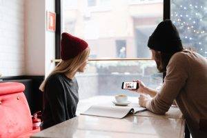 A man shows a photo on his smartphone to a woman sitting at a cafe table with a coffee cup and newspaper.