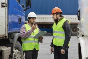Two men wearing safety vests and hard hats engage in discussion at a worksite, one holding a clipboard.