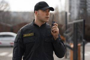 A security guard in a black uniform and cap holds a walkie-talkie to his mouth while standing outdoors.