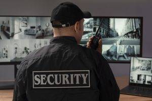 A security guard in uniform with 'SECURITY' on the back holds a walkie-talkie while monitoring multiple surveillance screens.