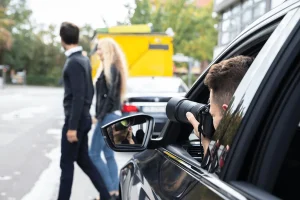 A private investigator photographing a man and woman walking by on the street from inside a car. The investigator's reflection is visible in the side mirror.