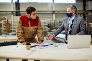 Two men wearing face masks—one in a suit and the other in work overalls—discussing project plans at a workspace with a laptop and coffee cup.