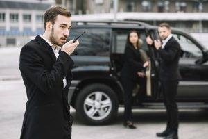 Three security agents in suits near a black SUV; one man speaks into a walkie-talkie while two others stand alert by the car.