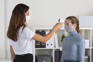 A woman in a white mask checks another woman's temperature with a handheld infrared thermometer while both wear face masks.