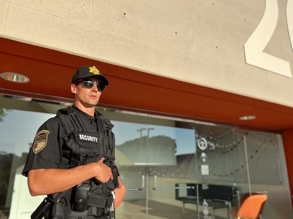 Armed security officer wearing sunglasses and a tactical vest with a security badge outside a building.