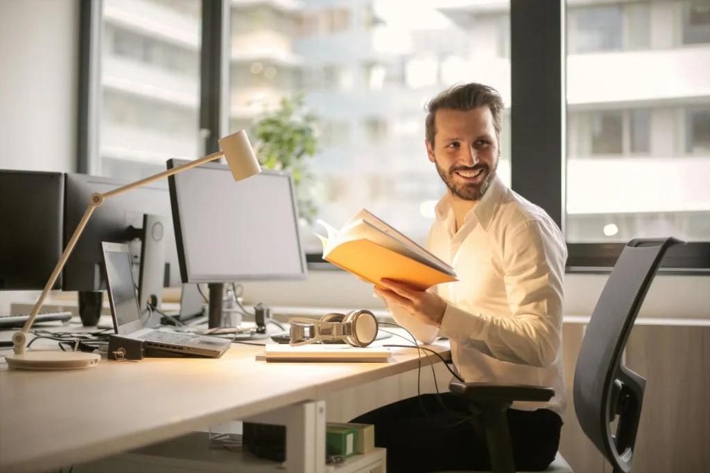 A man with a light shirt holding an orange book while sitting at a desk in an office environment.