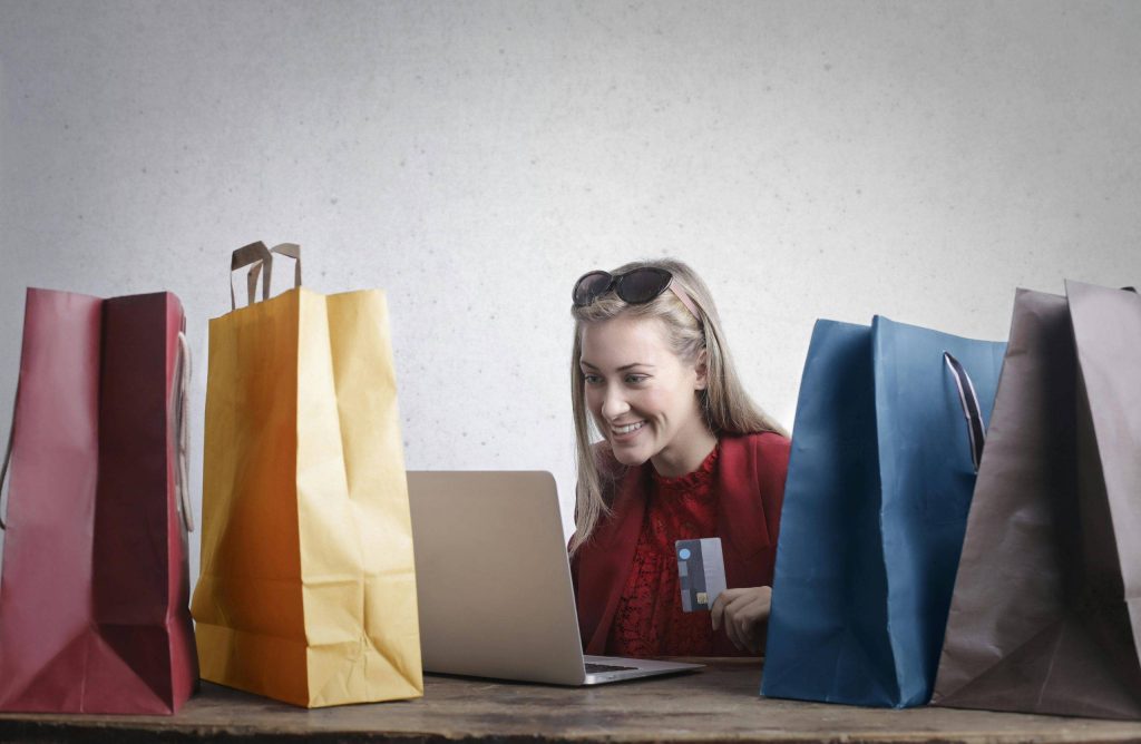 A woman smiling while shopping online with a laptop and colorful shopping bags around her.