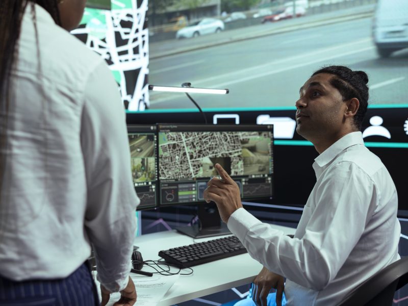 A man sitting at a desk with multiple monitors displaying maps, surveillance footage, and digital forensics data talks to a standing colleague in an office with large security screens in the background.