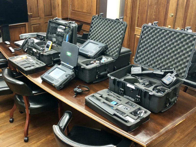 A wooden table in an office holds several open black equipment cases with electronic devices, screens, cables, and tools used for security; black chairs surround the table.