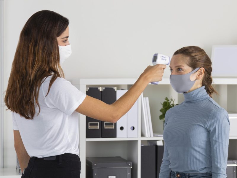 A woman in a white mask checks another woman’s temperature with a handheld infrared thermometer in an office, demonstrating executive protection protocols. Both women are wearing face masks.