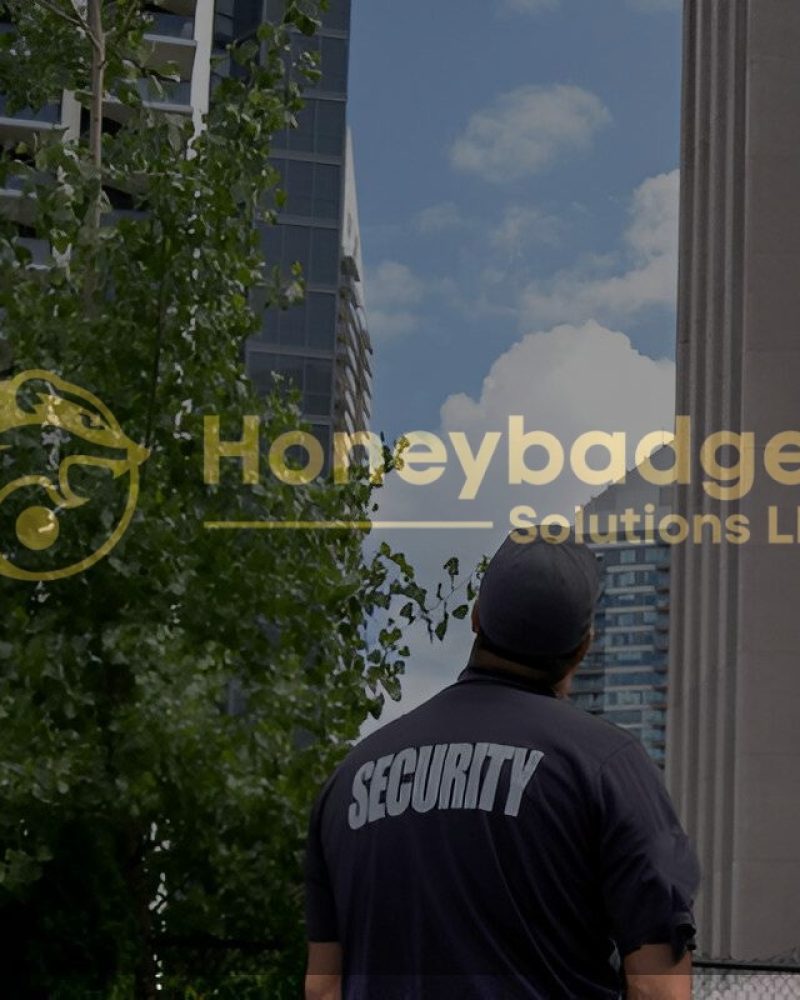 A security guard wearing a uniform stands outdoors, facing tall modern apartment buildings under a blue sky with clouds.